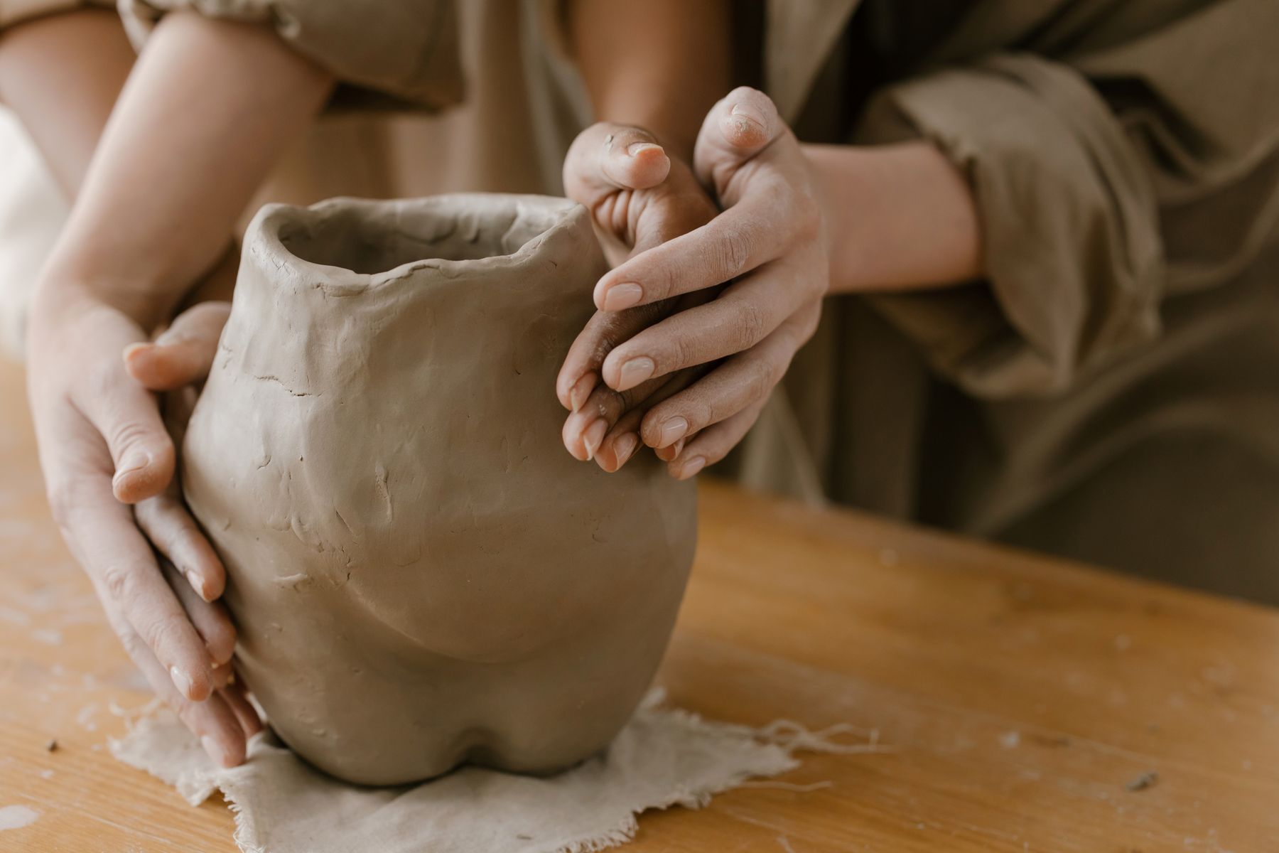 Hands shaping a clay pot together at a wooden worktable